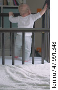 Curious baby stands in the crib, holding on to the edge, dressed in gray pants and a white shirt. The toddler takes his first steps with support. Focus on the crib railing. Стоковое видео, видеограф Воловодов Владимир / Фотобанк Лори