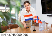 Positive young woman putting flag of Holland on the table with flag of Norway in conference room. Стоковое фото, фотограф Яков Филимонов / Фотобанк Лори