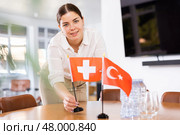 Positive young woman putting flag of Switzerland on the table with flag of Turkey in conference room. Стоковое фото, фотограф Яков Филимонов / Фотобанк Лори