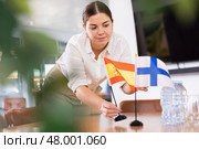 Positive young woman putting flag of Spain on the table with flag of Finland in conference room. Стоковое фото, фотограф Яков Филимонов / Фотобанк Лори