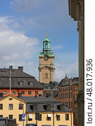Stockholm.Sweden.  Вид на собор Святого Николая с площади Birger Jarls Torg.  Светлый фасад Стокгольмского кафедрального собора (Storkyrkan) с зелёной башней и часами возвышается над крышами старого города. Снимок сделан с площади Birger Jarls Torg, на острове Риддархольмен. На переднем плане — исторические жилые здания в районе Гамла Стан с черепичными и жестяными крышами. Стоковое фото, фотограф Анна Чижова / Фотобанк Лори