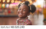 A joyful girl with curly hair grins widely, illuminated by the vibrant lights of a carnival. Стоковое AI-изображение, AI-художник Данила Большаков / Фотобанк Лори