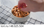 Hand picking an almond from a white ceramic bowl overflowing with almonds, resting on a checkered tablecloth atop a light wooden table, highlighting healthy snacking options. Стоковое видео, видеограф Ольга Ильина / Фотобанк Лори