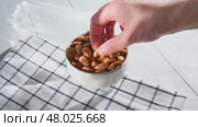 Hand picking an almond from a white ceramic bowl overflowing with almonds, resting on a checkered tablecloth atop a light wooden table, highlighting healthy snacking options. Стоковое видео, видеограф Ольга Ильина / Фотобанк Лори