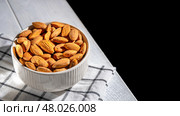 White bowl brimming with almonds resting on a checkered cloth atop a wooden table, creating a cozy, inviting atmosphere against a dark background. Perfect for highlighting healthy snacking. Стоковое фото, фотограф Ольга Ильина / Фотобанк Лори