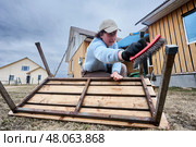 Cleaning metal frame of garden bench with wire brush, mature woman prepares surface for restoration work near wooden houses on cloudy day. Стоковое фото, фотограф Владимир Григорьев / Фотобанк Лори