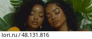 Two women with curly hair pose closely with their eyes closed, radiating serenity and grace amidst vibrant green plants in soft lighting. Стоковое AI-изображение, AI-художник Данила Большаков / Фотобанк Лори