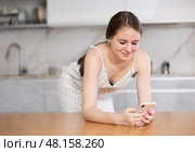 In the kitchen, teen woman leans on table with phone in hand, propping up chin with fist. Стоковое фото, фотограф Яков Филимонов / Фотобанк Лори