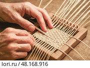 Skilled artisan weaving natural raffia strands on tabletop wooden loom creating eco friendly handcrafted textile for sustainable home craft decor market. Стоковое AI-изображение, AI-художник Павел Муравьев / Фотобанк Лори