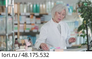 Mature female pharmacist standing behind the counter and working on computer, she checks the availability of medical medicines in a spreadsheet and prepares to collect an online order. Стоковое видео, видеограф Яков Филимонов / Фотобанк Лори
