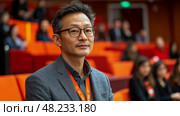 A thoughtful man wearing glasses sits in an auditorium, observing a conference, while attendees around him participate in discussions. Стоковое AI-изображение, AI-художник Данила Большаков / Фотобанк Лори