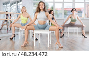 Attractive young woman blending sensuality with fashion while practicing seductive movements sitting on chair in group high heels dance class with sports women at dance studio. Стоковое фото, фотограф Яков Филимонов / Фотобанк Лори
