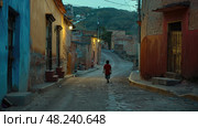 A young boy walks down a narrow cobblestone street in a village at dusk, the buildings on either side cast long shadows. Стоковое AI-изображение, AI-художник Данила Большаков / Фотобанк Лори
