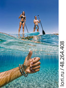 Split shot image taken below the wavy water surface. Thumbs up male hand in bracelet is underwater, two adults are on boards in the foreground. Стоковое AI-изображение, AI-художник Катерина Ковалева / Фотобанк Лори