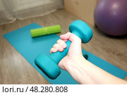 Бирюзовая гантель в женской руке на фоне фитнес-коврика, фитбола и рола в комнате. Упражнение для мышц рук. Стоковое фото, фотограф Юлия Королева / Фотобанк Лори