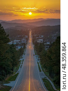 A golden sunset casts warm light over a long road stretching into the distance near a town, framed by mountains and trees on either side. Стоковое AI-изображение, AI-художник Данила Большаков / Фотобанк Лори