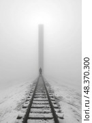 A person strides along icy railway tracks shrouded in thick fog, heading towards a striking vertical structure in the distance. Стоковое AI-изображение, AI-художник Данила Большаков / Фотобанк Лори