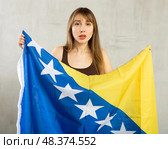 Sad young woman holding flag of Bosnia and Herzegovina against unicoloured background. Стоковое фото, фотограф Яков Филимонов / Фотобанк Лори