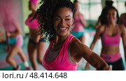A woman with curly hair smiles and leads a Zumba class in a brightly lit fitness studio. Стоковое AI-изображение, AI-художник Данила Большаков / Фотобанк Лори