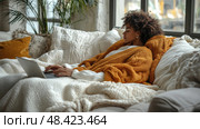 A woman enjoys a comfortable afternoon on the couch, engaged in her laptop work while surrounded by cozy blankets and fluffy pillows. Стоковое AI-изображение, AI-художник Данила Большаков / Фотобанк Лори