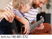 Handsome dad teaches his cute son to play the guitar sitting on sofa in the living room at home. Happy Father's Day. Father teaching his child to play guitar. Kid Learning music skill. Art education. Стоковое фото, фотограф Марина Демидюк / Фотобанк Лори