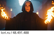 An older woman in a black hooded cloak conjures flames in her hands inside a mystical dark alley. Стоковое AI-изображение, AI-художник Igor Tichonow / Фотобанк Лори
