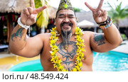 A tattooed Polynesian man with a floral lei smiling and gesturing peace signs at a cultural event. Стоковое AI-изображение, AI-художник Данила Большаков / Фотобанк Лори