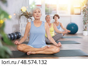 Elderly woman in company of hatha yoga lovers performs half-lotus pose, Ardha Padmasana. Стоковое фото, фотограф Яков Филимонов / Фотобанк Лори