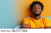 A confident young man with stylish hair sits on a couch, arms crossed, against a vibrant blue and orange wall. Стоковое AI-изображение, AI-художник Данила Большаков / Фотобанк Лори