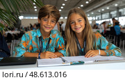 Two joyful kids dressed in vibrant matching shirts engage in schoolwork at a lively educational event, surrounded by other participants. Стоковое AI-изображение, AI-художник Данила Большаков / Фотобанк Лори
