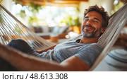 A man smiles contentedly while lounging in a hammock, basking in the warmth of a bright afternoon indoors among lush plants. Стоковое AI-изображение, AI-художник Данила Большаков / Фотобанк Лори