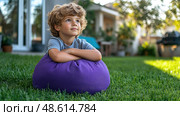 A young boy rests comfortably on a purple beanbag chair in a sunny backyard, gazing thoughtfully into the distance during a peaceful afternoon. Стоковое AI-изображение, AI-художник Данила Большаков / Фотобанк Лори