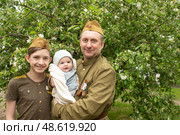 Отец и двое его сыновей празднуют 9 мая, День Победы. Мужчина и подросток одеты в одежду, стилизованную под форму солдат Советской Армии. Младенец на руках у отца. Стоковое фото, фотограф Яна Проничева / Фотобанк Лори