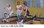 Two health conscious women of different ages perform a Pilates exercise using a reformer bed, where a female instructor helps them do it correctly. Стоковое видео, видеограф Яков Филимонов / Фотобанк Лори