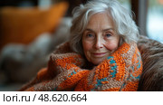 An elderly woman enjoys a quiet evening indoors, wrapped in a colorful knitted blanket, exuding warmth and contentment. Стоковое AI-изображение, AI-художник Данила Большаков / Фотобанк Лори