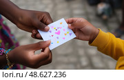 Two hands exchanging a colorful card with hearts and shapes in a vibrant community setting. Стоковое AI-изображение, AI-художник Сергей Толмачёв / Фотобанк Лори
