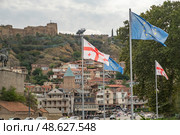 Flag display in Tbilisi with historical fortress and hillside homes visible in background during an overcast day. Редакционное фото, фотограф Климов Денис / Фотобанк Лори