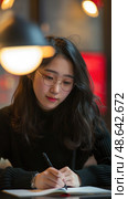 A young woman wearing glasses focuses on writing in a notebook at a café, surrounded by warm, inviting lights during the evening hours. Стоковое AI-изображение, AI-художник Данила Большаков / Фотобанк Лори