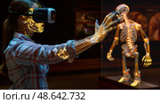 A young woman, wearing a virtual reality headset, reaches out to engage with a glowing holographic skeleton displayed in an interactive museum exhibit. Стоковое AI-изображение, AI-художник Данила Большаков / Фотобанк Лори