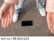 A person stands with outstretched hands, preparing to lift a smartphone from the pavement, indicating a moment of concern outdoors. Стоковое фото, фотограф Данила Большаков / Фотобанк Лори