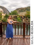 A woman stands on a wooden deck, wearing a blue dress, admiring the peaceful lake and picturesque mountains in the background under a cloudy sky, Главрыба, Республика Дагестан, Казбековский район, село Ново-Зубутли. Стоковое фото, фотограф Данила Большаков / Фотобанк Лори