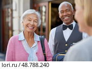 Senior woman arriving at hotel greeted by smiling porter and concierge. Стоковое AI-изображение, AI-художник Юрий Поздников / Фотобанк Лори