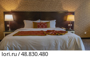 The double bed is covered with white sheets. The red bedspread is neatly and beautifully laid. Pillows at the headboard. The bedside lamps are lit. Golden beige patterned wallpaper on the wall. Стоковое фото, фотограф Вера Ивановна Кубарева / Фотобанк Лори