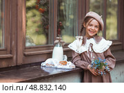 Vintage accessories and retro-style clothes. A small, beautiful girl with bows in her hair, in a beige dress and a beret outdoors with bagels and milk in a string bag. Стоковое фото, фотограф Photojuli / Фотобанк Лори
