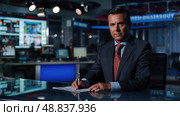A man in a suit and tie sits at a desk in a television studio, writing notes on a piece of paper. He looks focused and serious, likely preparing for a news broadcast. Стоковое AI-изображение, AI-художник Данила Большаков / Фотобанк Лори