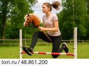 Hobby horsing. Focused young woman clearing small equestrian style hurdle while racing hobby horse across lush meadow, unique alternative sport concept for leisure communities. Стоковое AI-изображение, AI-художник Павел Муравьев / Фотобанк Лори