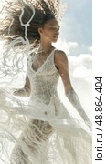 A woman with curly hair is wearing a white lace dress with a plunging neckline. The dress is flowing in the wind. The woman is standing in a field on a sunny day. Стоковое AI-изображение, AI-художник Данила Большаков / Фотобанк Лори