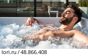 A bearded man relaxes in a hot tub filled with ice on a sunny day. He is enjoying the cold therapy, which can be beneficial for recovery and muscle soreness. Стоковое AI-изображение, AI-художник Данила Большаков / Фотобанк Лори