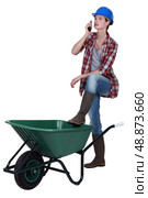 Tradeswoman speaking into a walkie-talkie and propping her foot up on a wheelbarrow. Стоковое фото, агентство Ingram Publishing / Фотобанк Лори