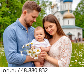 A man and a woman holding a baby in front of an Orthodox church. Since 2008, Russia has celebrated July 8 as the Day of Family, Love, and Fidelity, dedicated to Peter and Fevronia of Murom. Стоковое AI-изображение, AI-художник Сергей Елизарьев / Фотобанк Лори